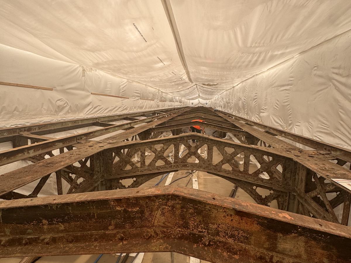 Vue intérieure d'un confinement sous bâche tendue enveloppant une charpente métallique rivetée à peinture écaillée, avant décapage par projection d'abrasif.
