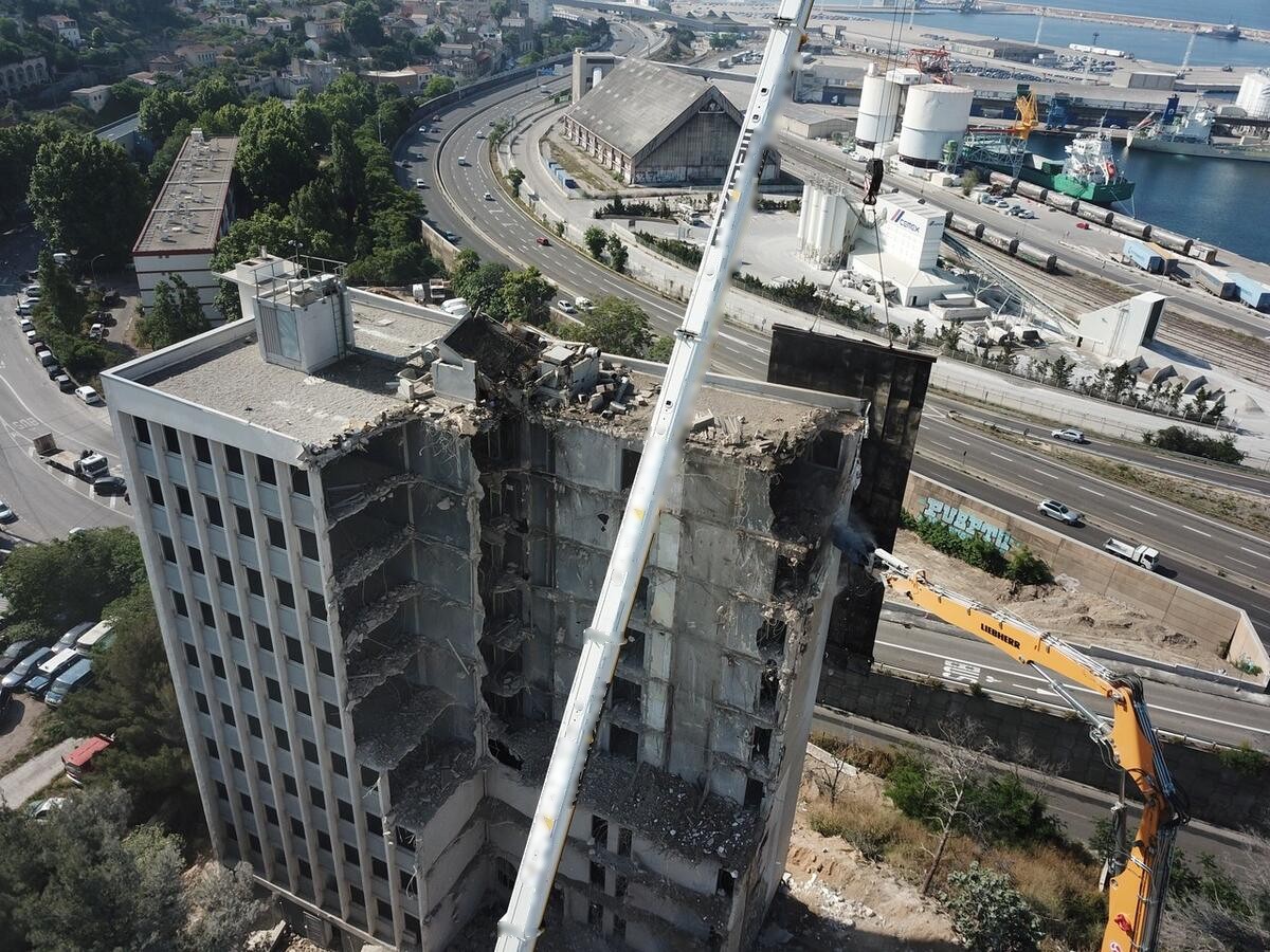 Vue aérienne d'un immeuble de grande hauteur en cours de démolition, pelle à grand bras et grue treillis, entre une voie rapide et une zone portuaire.