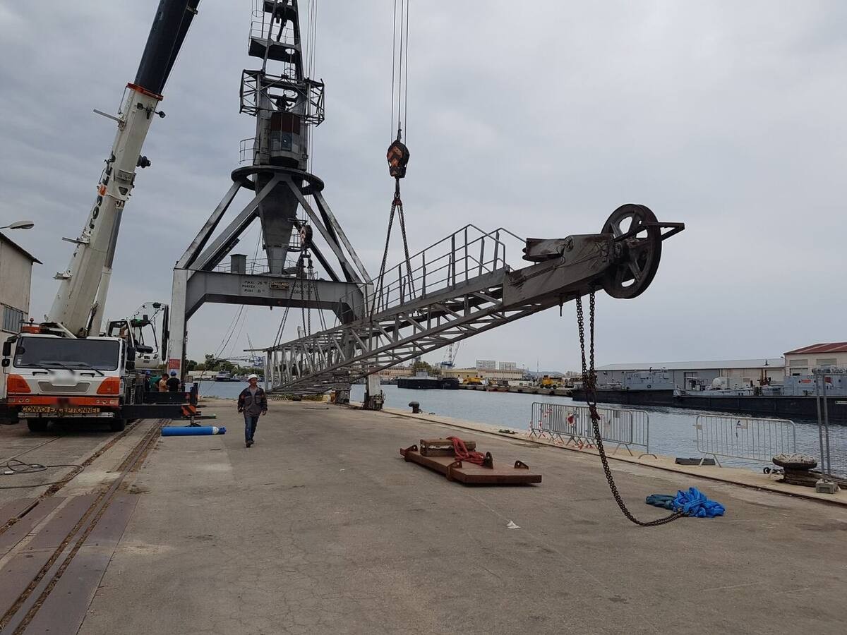 Flèche en treillis d'une grue portuaire déposée sur un quai, élinguée à une grue mobile, en cours de démontage par oxycoupage.
