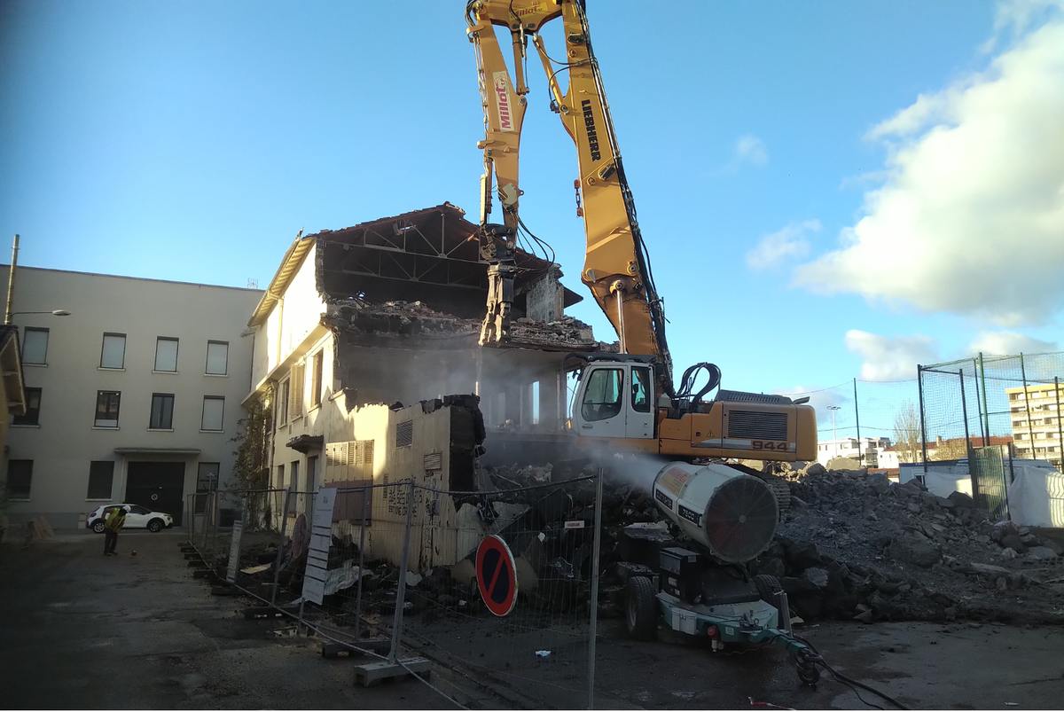 Pelle de démolition à grand bras équipée d'une pince hydraulique attaquant la charpente d'un bâtiment, panache de poussière rabattu par un canon brumisateur au premier plan.